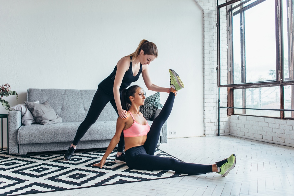 Fit woman doing stretching exercises with the help of friend holding her leg at home.