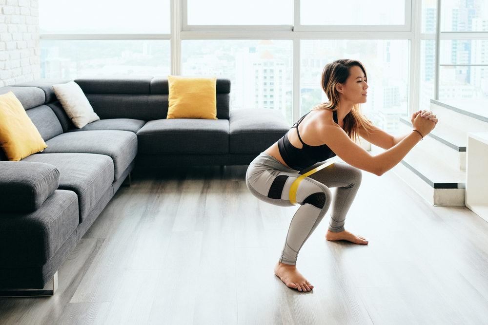 Fit young Pacific Islander woman training at home. Beautiful female athlete working out for wellbeing in domestic gym, training legs muscles doing side to side squats with elastic band.